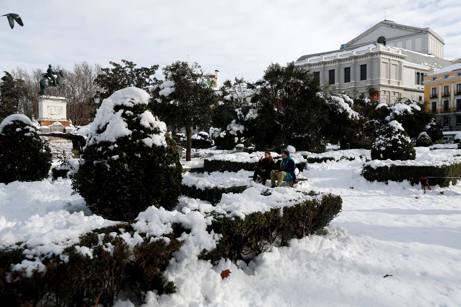 Men sit on a bench in a snow-covered square with the Royal Theatre in the background, in Madrid