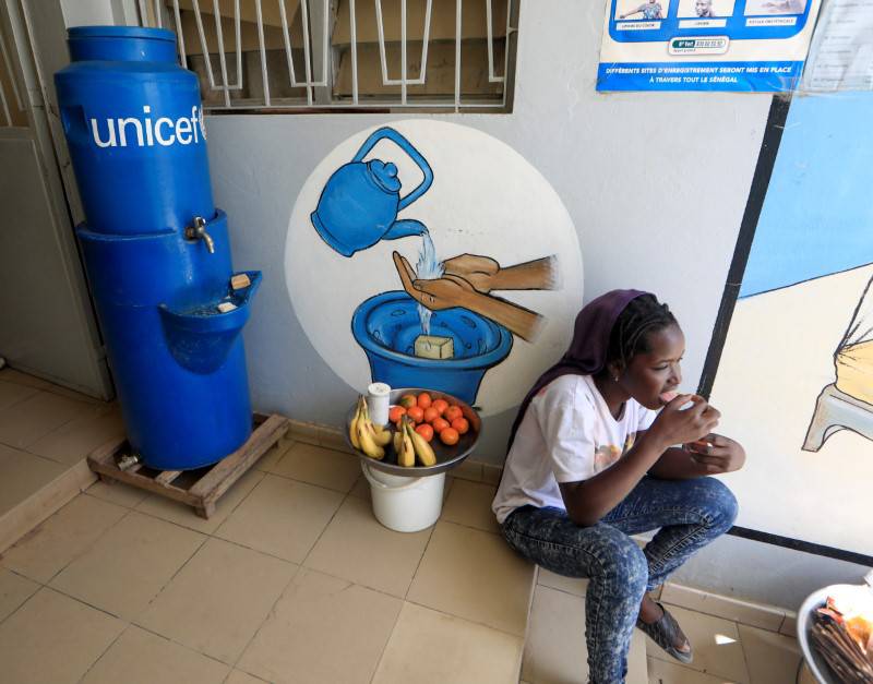 A girl eats as she sits next to a handwashing station, while she sells fruits at the entrance of a medical center in Pikine, on the outskirts of Dakar