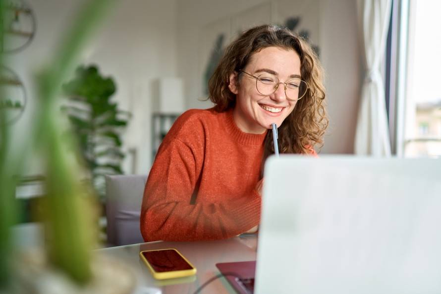 Happy,Young,Woman,Using,Laptop,Sitting,At,Desk,Writing,Notes