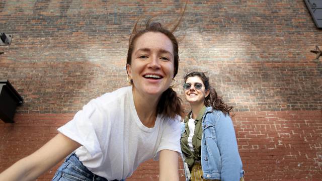 Portrait of two young millennial women, wearing casual clothing, move playfully in front of a brick wall.