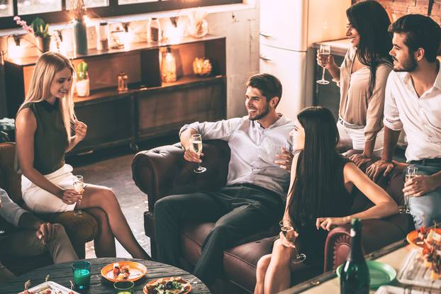 Carefree time with friends. Top view of cheerful young people enjoying food and drinks while spending nice time in cofortable chairs at home together