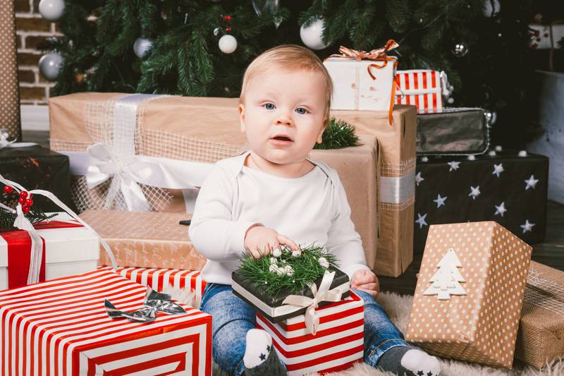 theme winter and Christmas holidays. Child boy Caucasian blond 1 year old sitting home floor near Christmas tree with New Year decor on shaggy carpet skin receives gifts, opens gift boxes in evening