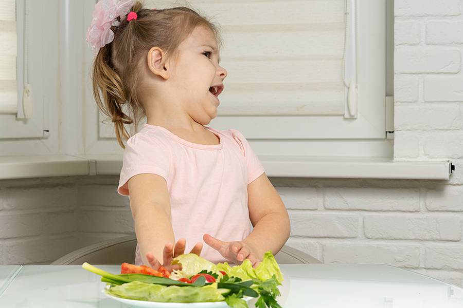 A Little Girl Sits At A Table In The Kitchen And Pushes Away A P