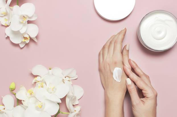 Young woman moisturizes her hand with cosmetic cream lotion opened container with cream body milk White Phalaenopsis orchid flowers on pink background Flat lay top view minimalism style Beauty concept