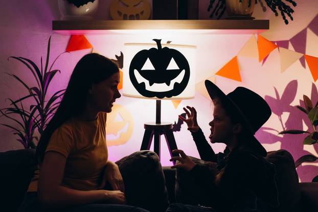 Silhouette of young mother sitting on couch in child room with cute little daughter tells spooky Halloween stories. Funny mom and girl kid have fun at home. Celebration and Autumn holiday concept