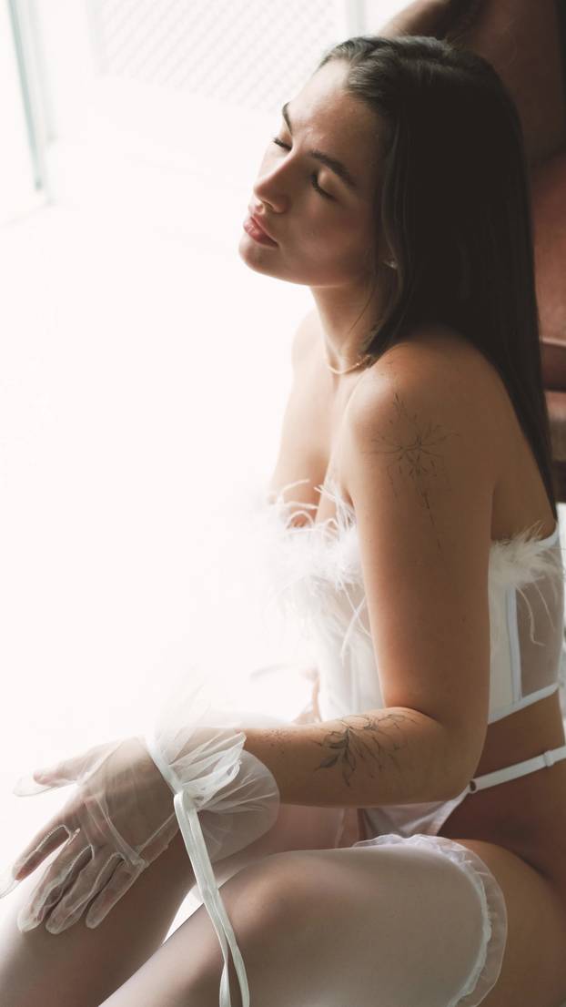 Portrait of young beautiful model posing in white lace lingerie, corset and stockings near a large window. Calm cute brunette with long hair in fashionable lingerie.