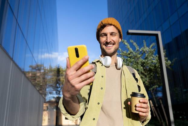 Smiling,Young,Man,Using,Phone,And,Drinking,Coffee,Outdoors,In