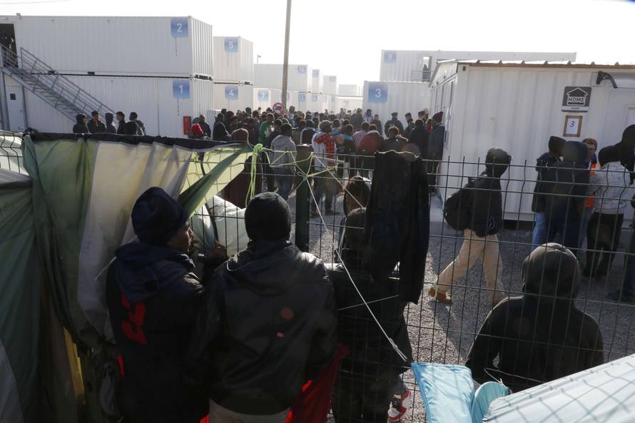A small group of migrants stand at a fence and look as other migrants gather by the containers used as temporary housing on the eve of the evacuation of the makeshift camp called the "Jungle" in Calais