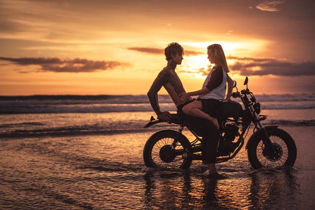 girlfriend touching shirtless boyfriend on motorbike at beach during sunset