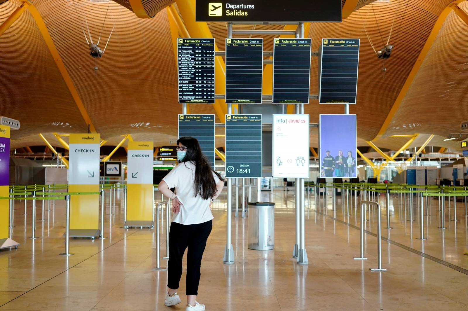 A woman wearing a protective mask stands in front of flight screens at the almost empty Madrid's Adolfo Suarez Barajas Airport