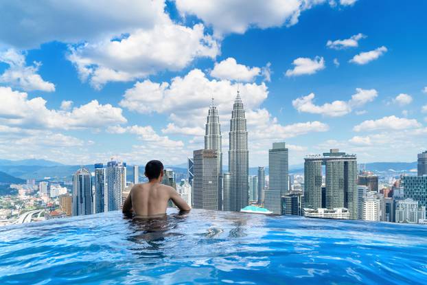 Back of tourist in a swimming pool on rooftop with Kuala Lumpur downtown view and blue sky. Malaysia travel trip in vacation and holidays concept in Asia. Skyscraper and high-rise buildings at noon.