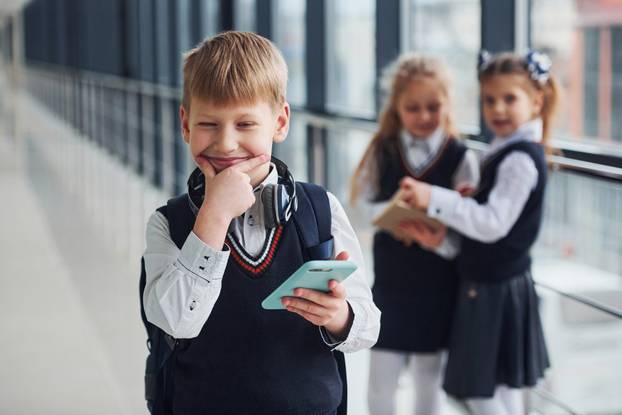 Little boywith phone and headphones standing if front of school kids in uniform that together in corridor. Conception of education