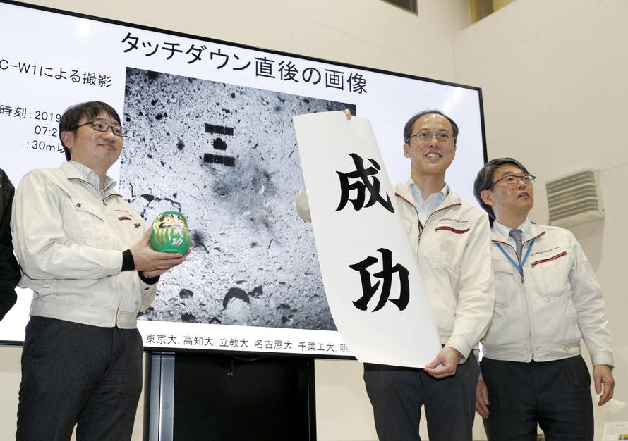 JAXA associate professor Yuichi Tsuda holds a banner reading 'success' in front of an image of the Hayabusa 2 space probe's landing on the Ryugu asteroid in Sagamihara
