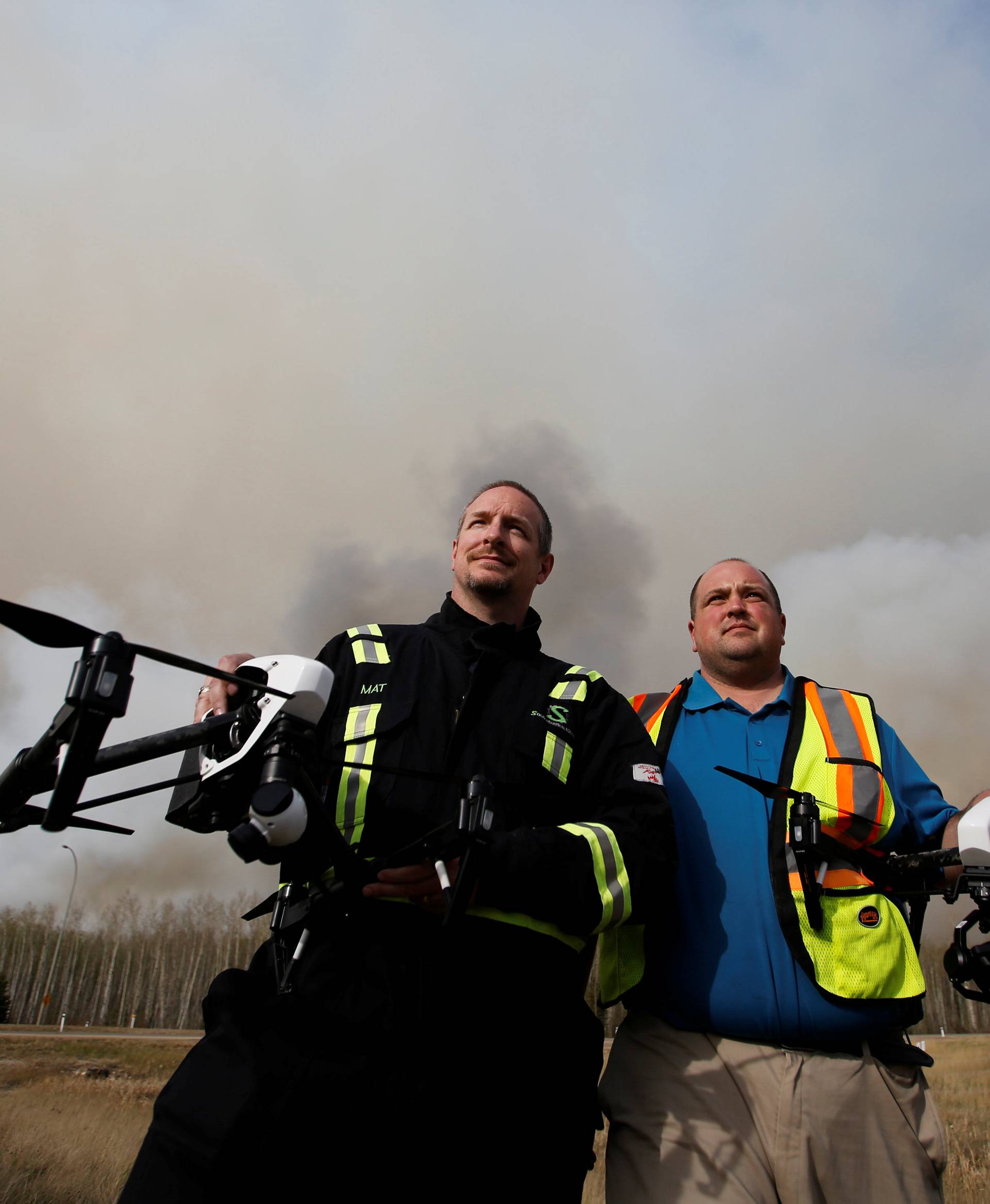 Mat Matthews, operations and safety manager with Elevated Robotic Services, and Ron Windmueller, owner of Droneology, pose with drones as smoke rises from wildfires near Fort McMurray, Alberta, Canada