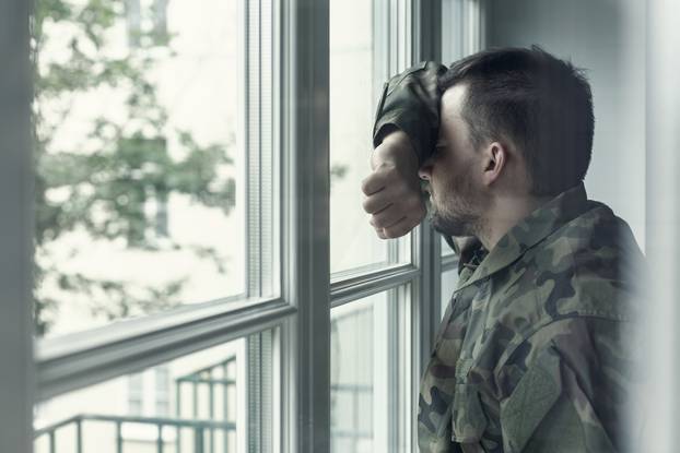 Depressed and sad soldier in green uniform with trauma after war standing near the window