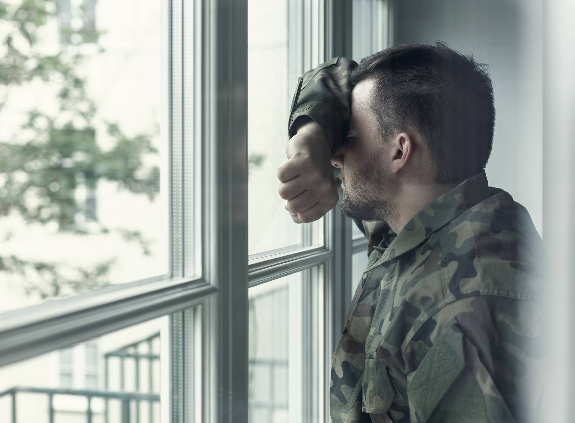 Depressed and sad soldier in green uniform with trauma after war standing near the window