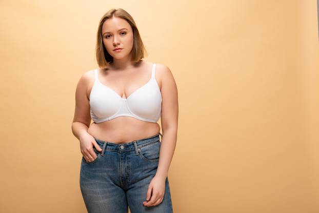 pensive overweight girl looking at camera while holding hand in pocket isolated on beige, body positivity concept