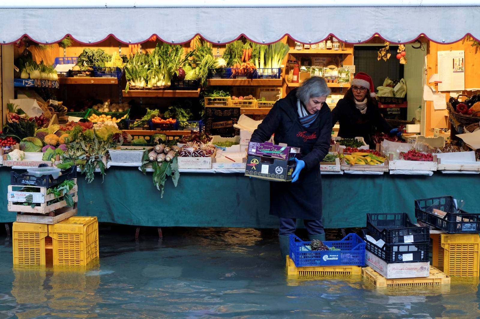 A woman carries a box in front of her vegetable stand during high tide in Venice