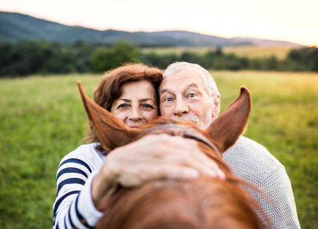 A crazy senior couple standing by a horse outside in nature, looking over his head.