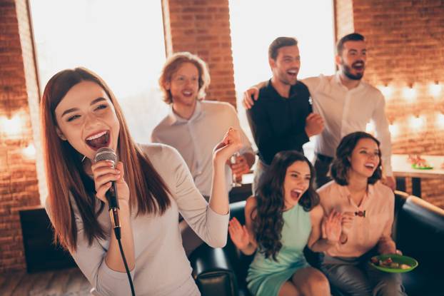 Close up photo best friends buddies karaoke gathering hang out sing she her ladies soloist he him his guys help yell shout scream words song wear dresses shirts formalwear sit sofa loft room indoors
