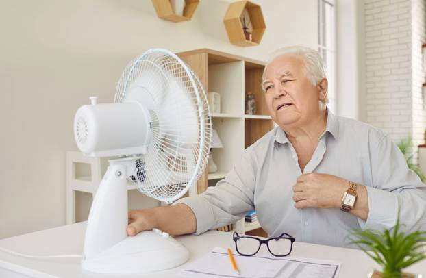 Tired upset senior retired person using electric fan during heatwave sitting at the desk.