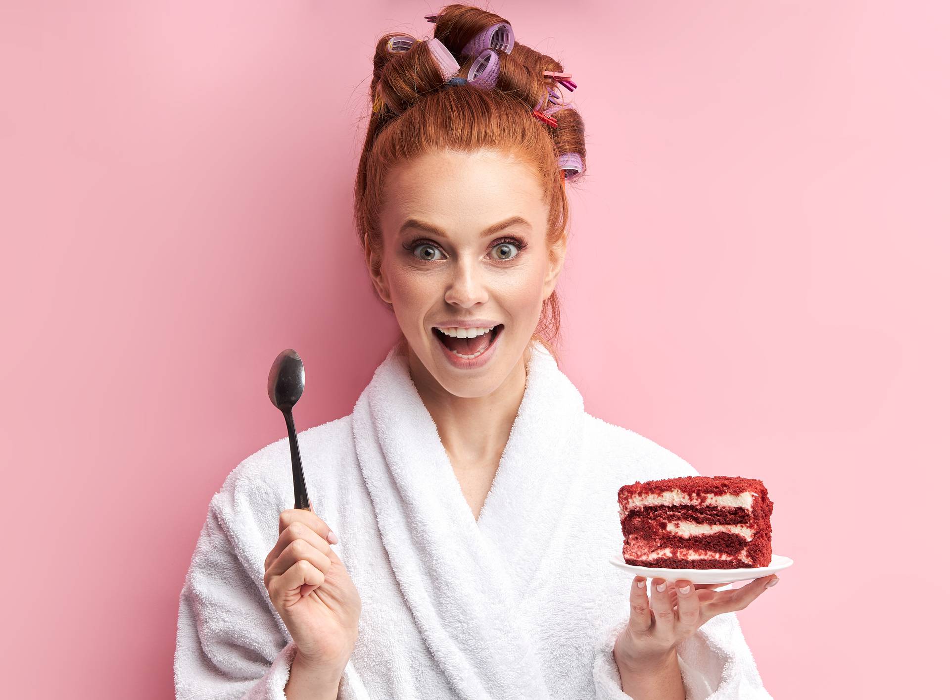 Happy young woman going to eat tasty cake on pink background