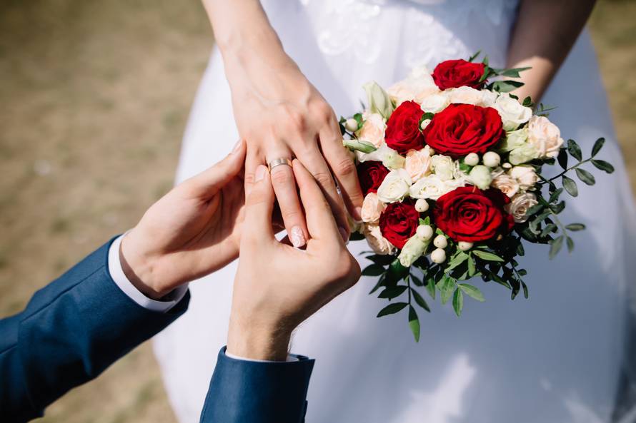 Love and marriage. Fine art rustic wedding ceremony outside. Groom putting golden ring on the bride's finger. Bouquet of red and white roses in hands
