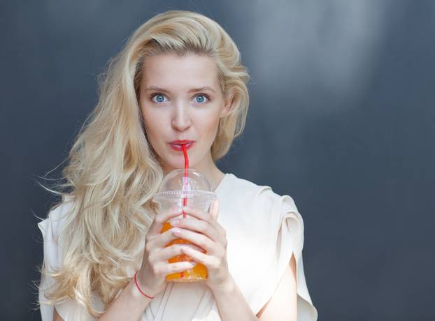 Beautiful sexy blonde with blue eyes drinking beverage through a straw on a hot summer day near the wall