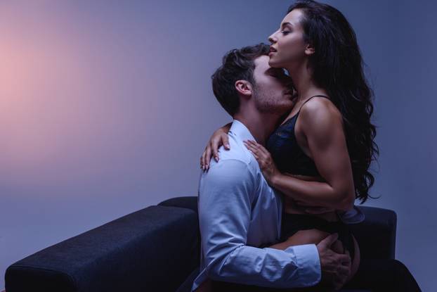 young man in white shirt hugging and kissing passionate girl in black lingerie while sitting on couch on purple background