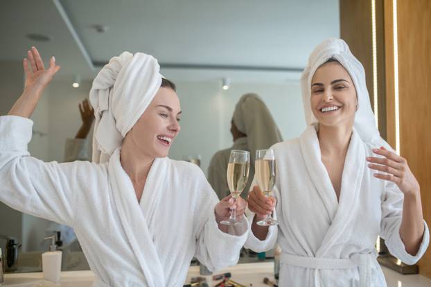 Two pretty girls in white robes drinking champagne and enjoying